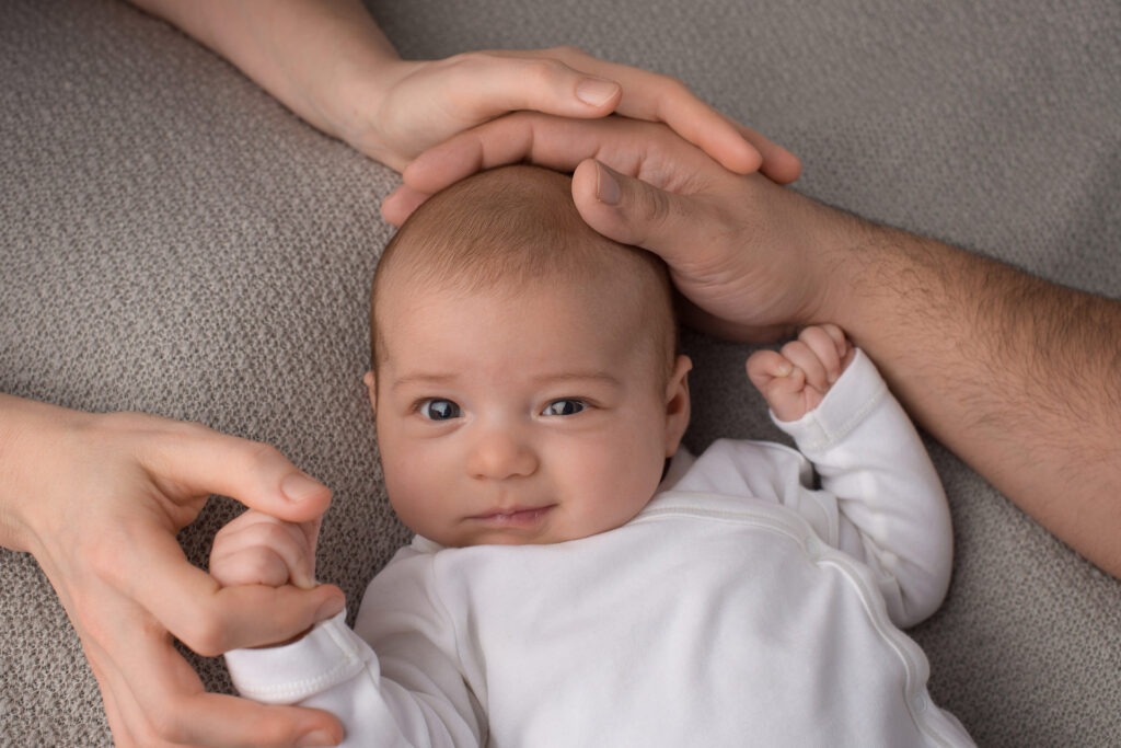 Baby liegt auf dem Rücken, Elternhände schützend am Kopf – Babyfotoshooting Ludwigsburg