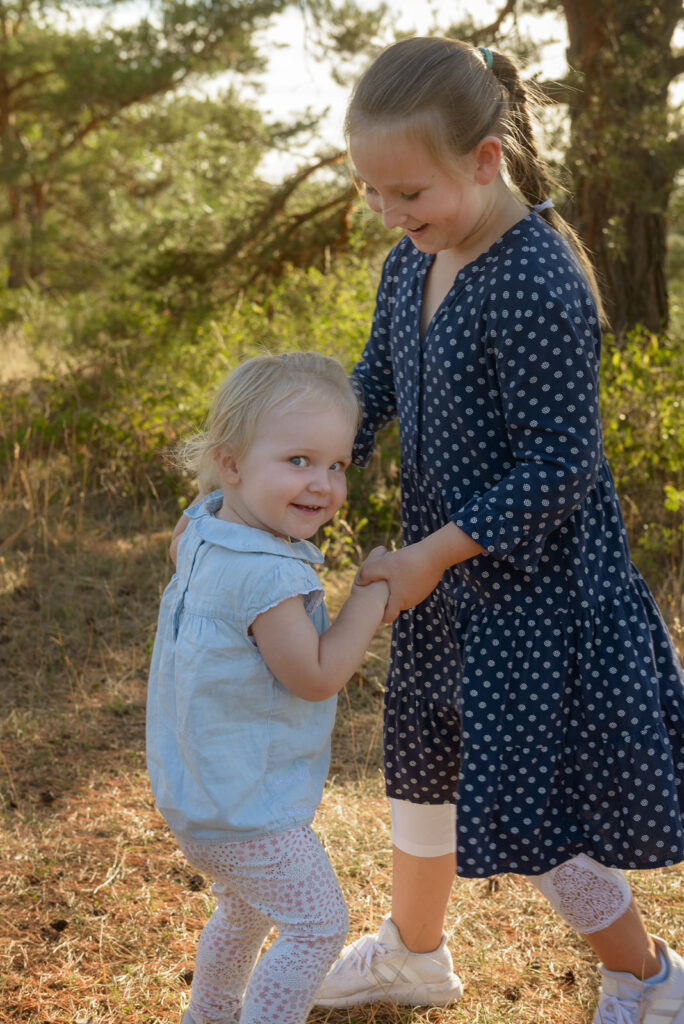 Ein verschmitzt lächelndes Kleinkind das mit seiner Schwester auf einer Wiese während des Fotoshootings. 