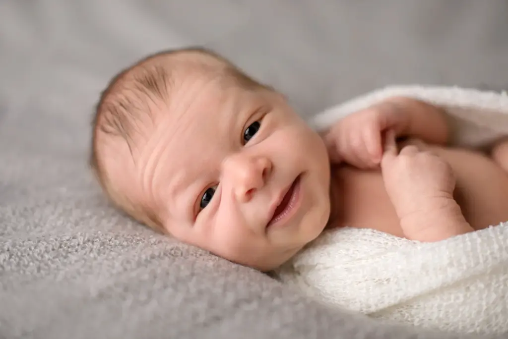 Freundlich lächelndes Neugeborenes, seitlich liegend auf weichem grauem Hintergrund – Babyfotografie Hemmingen